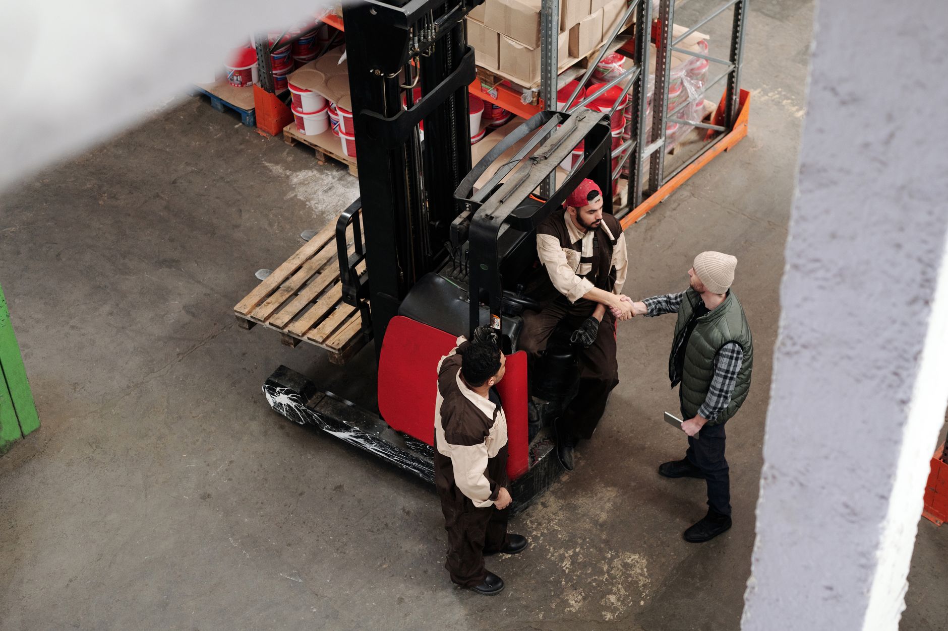 Three warehouse workers in conversation and handshake next to a forklift in an industrial setting.