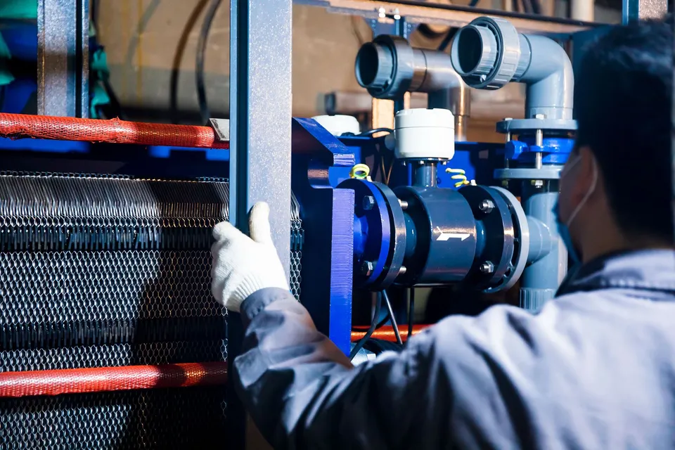 A worker in blue sleeves inspects machinery pipes and equipment at an industrial plant.